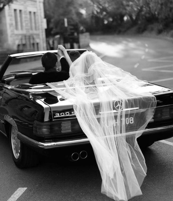 Schwarz-Weiß-Foto: Der Schleier einer Braut weht aus einem alten Mercedes