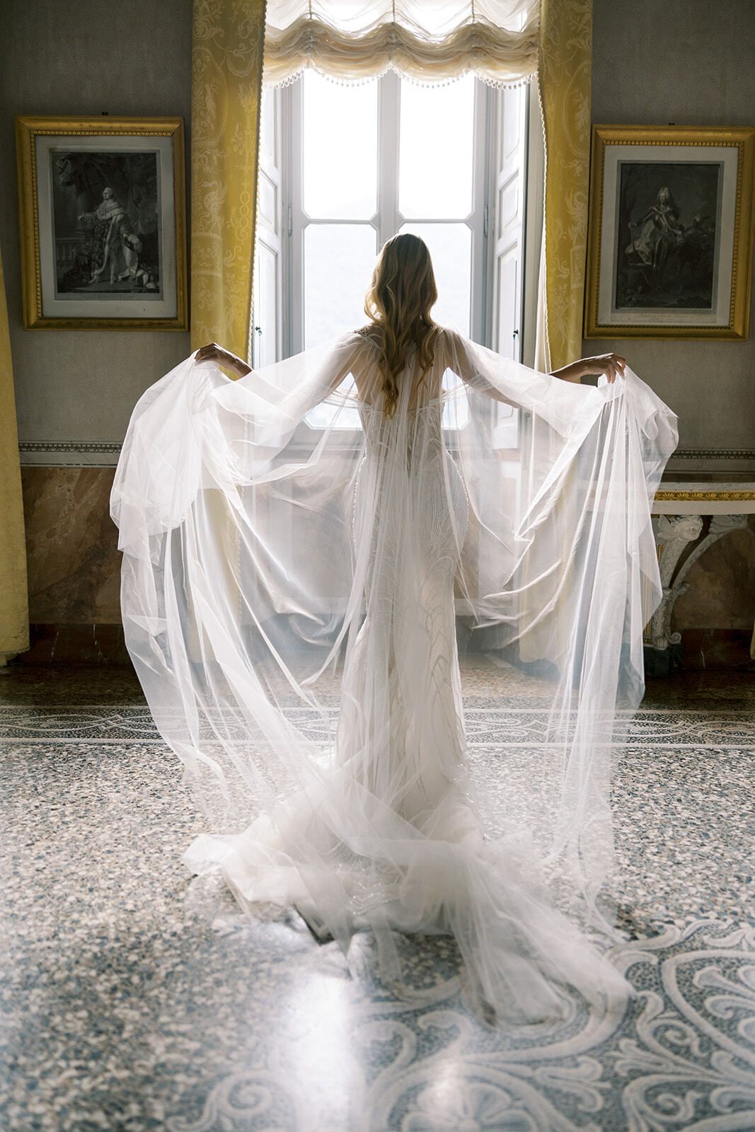 Bride with a sheer cape at a tall window in a villa salon