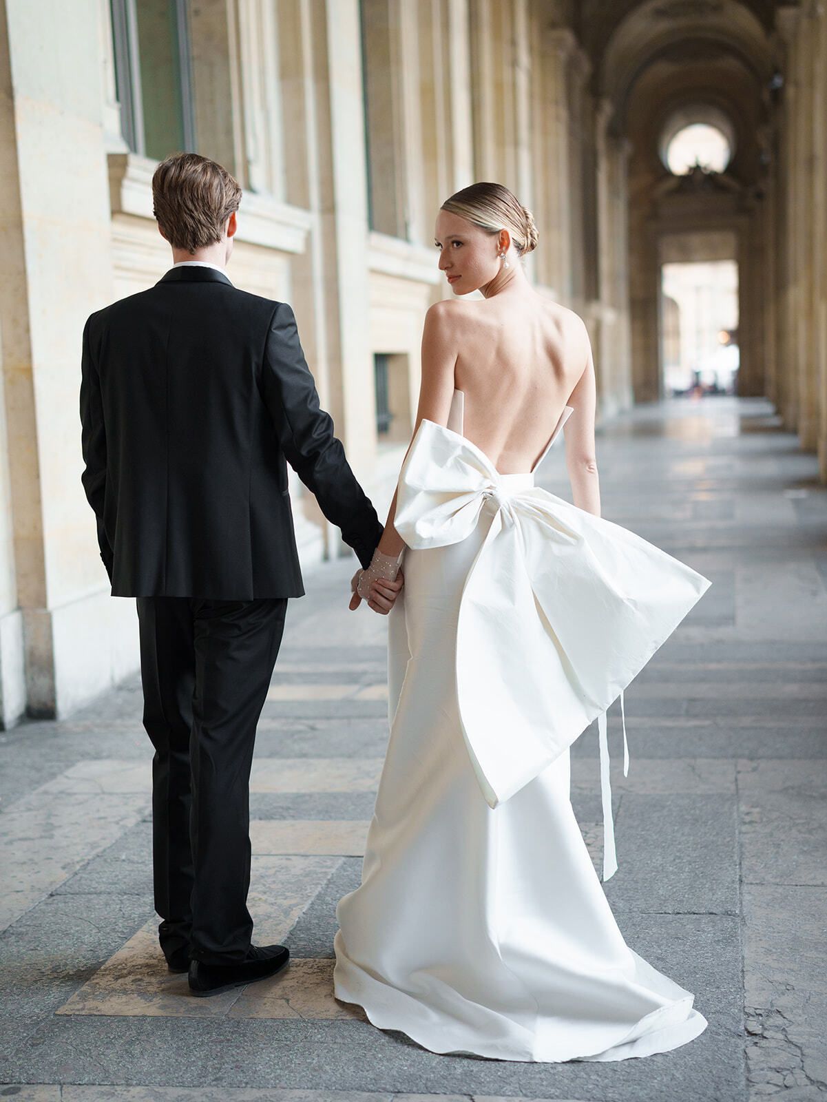 Braut im Kleid mit Rückenschleife geht mit dem Bräutigam durch die Arkaden des Louvre