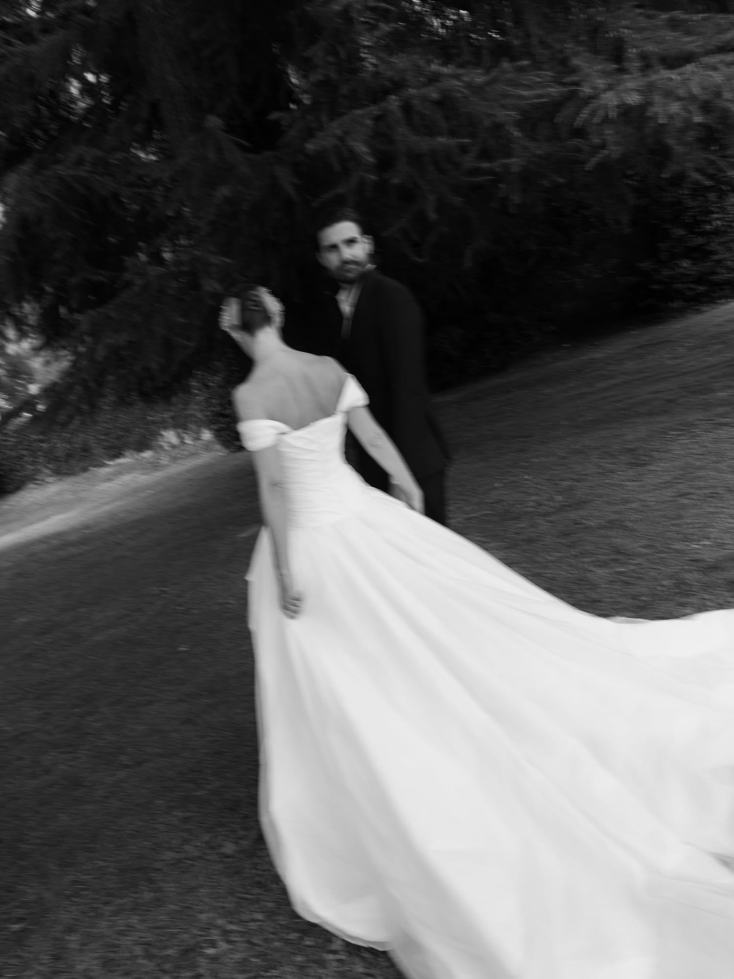 Black and white photo of a bride's long train on a garden path, groom ahead