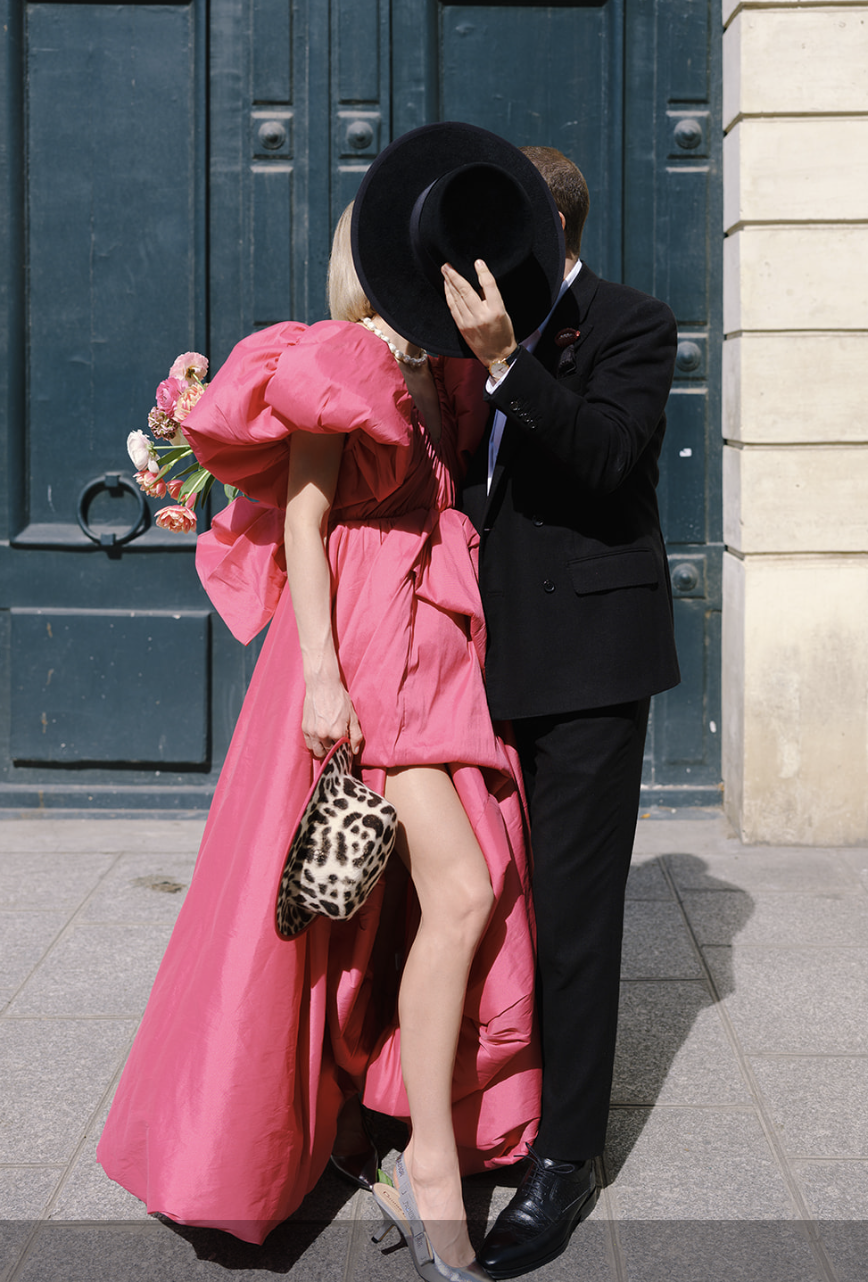 Mujer con vestido rosa besando a un hombre oculto tras un sombrero, París