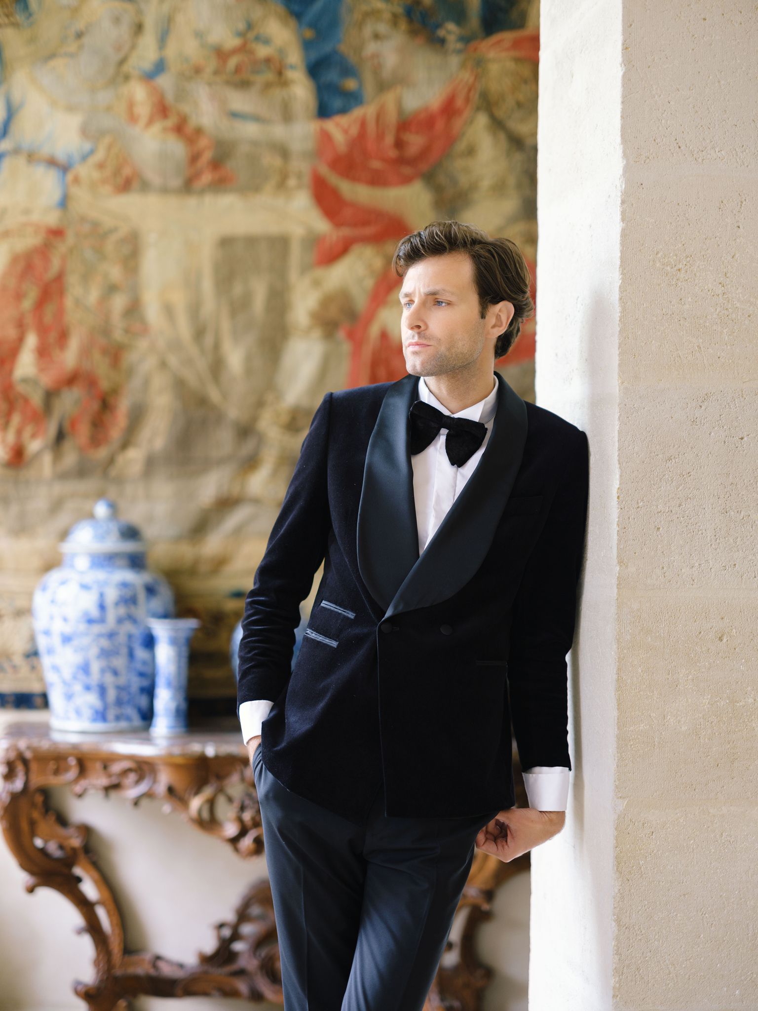 身着天鹅绒礼服外套的新郎站在 Château de Villette 的挂毯旁