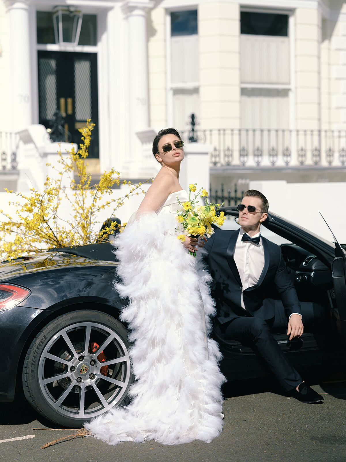 Braut in Federkleid und Sonnenbrille mit dem Bräutigam an einem Sportwagen