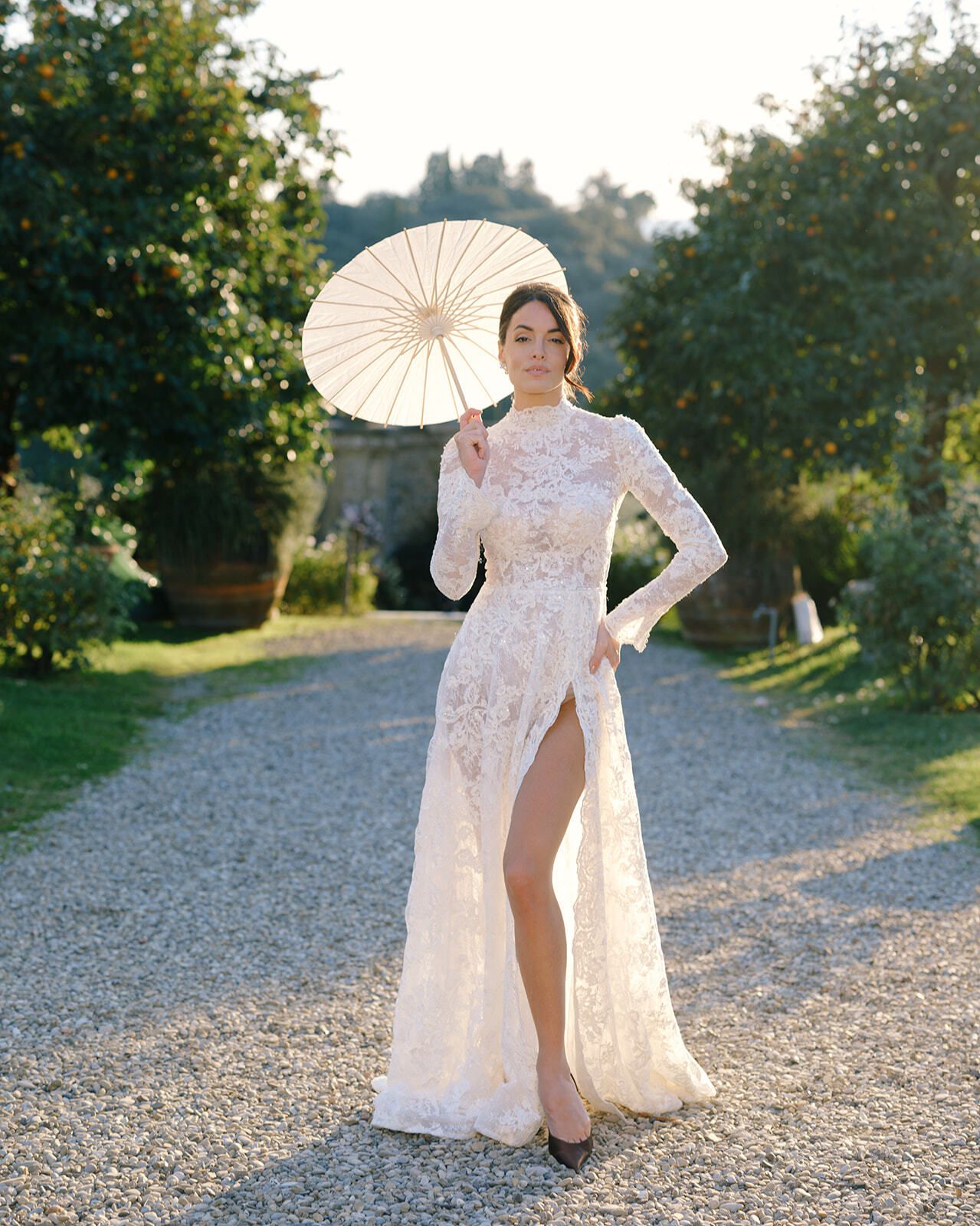 Novia con vestido de encaje y sombrilla en el jardín de la villa