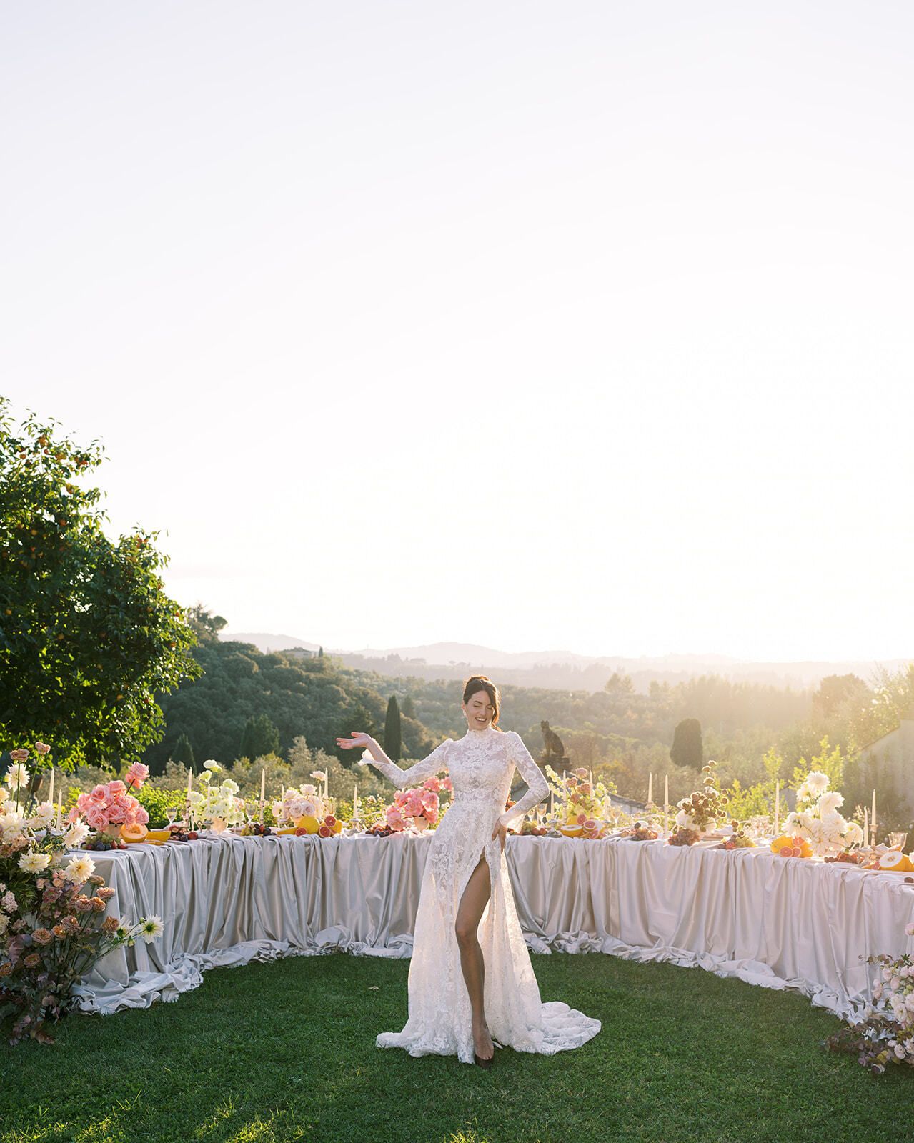 Novia junto a una mesa larga llena de fruta en el césped al atardecer, Toscana