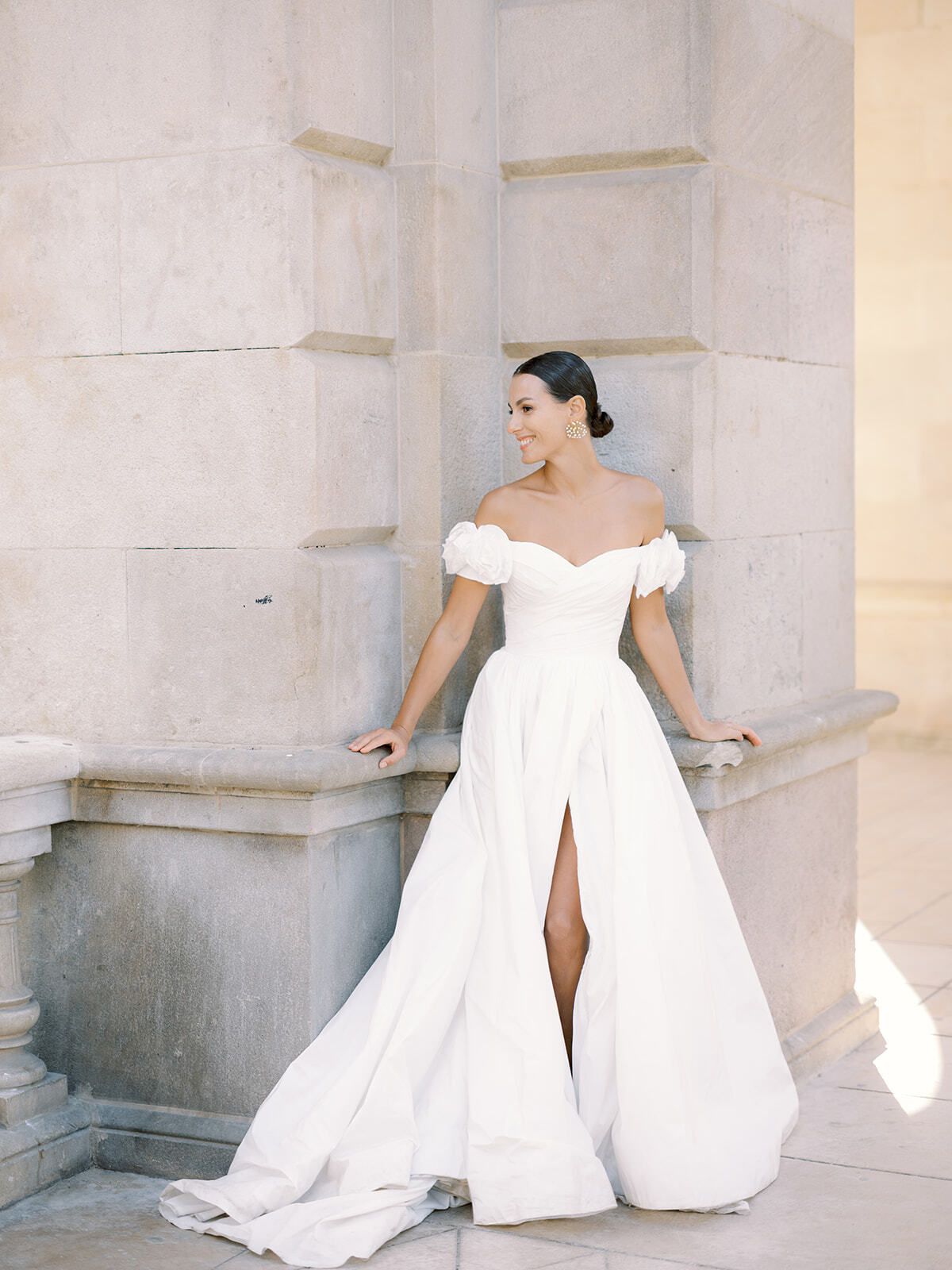 Novia con vestido de hombros descubiertos y abertura junto a una columna de piedra