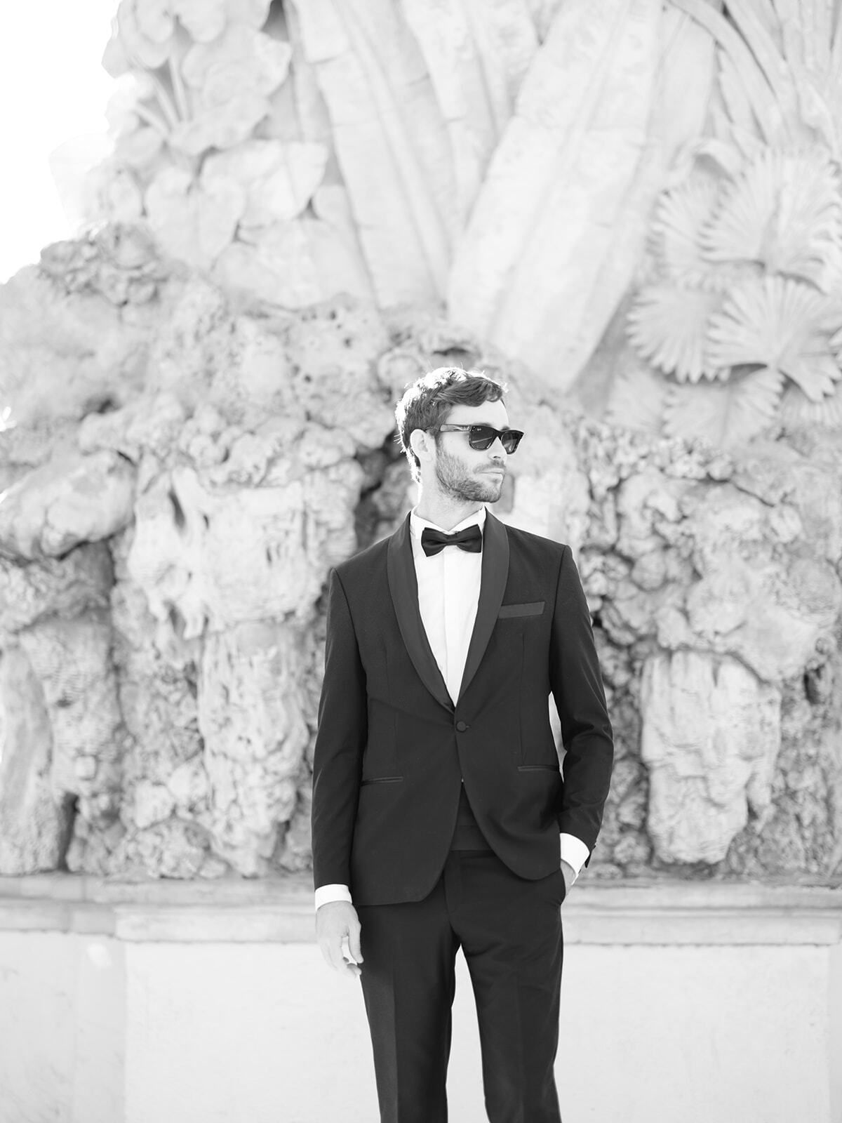 Retrato en blanco y negro de un novio con gafas de sol ante una fuente esculpida