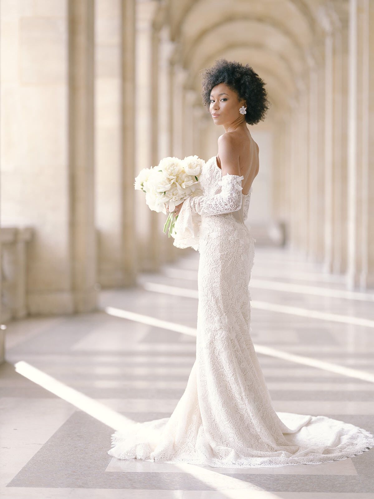 Braut im Spitzenkleid mit weißem Strauß unter den Arkaden des Louvre