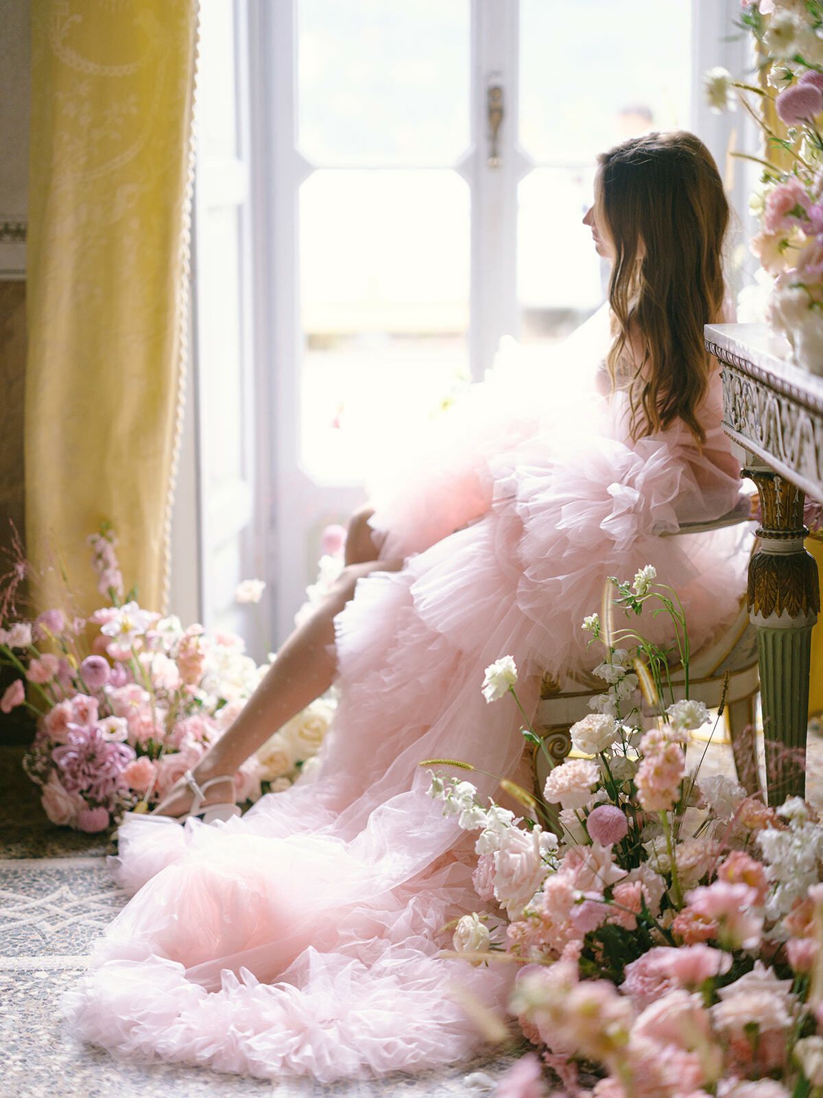 Novia con vestido rosa empolvado en el alféizar de una ventana rodeada de flores