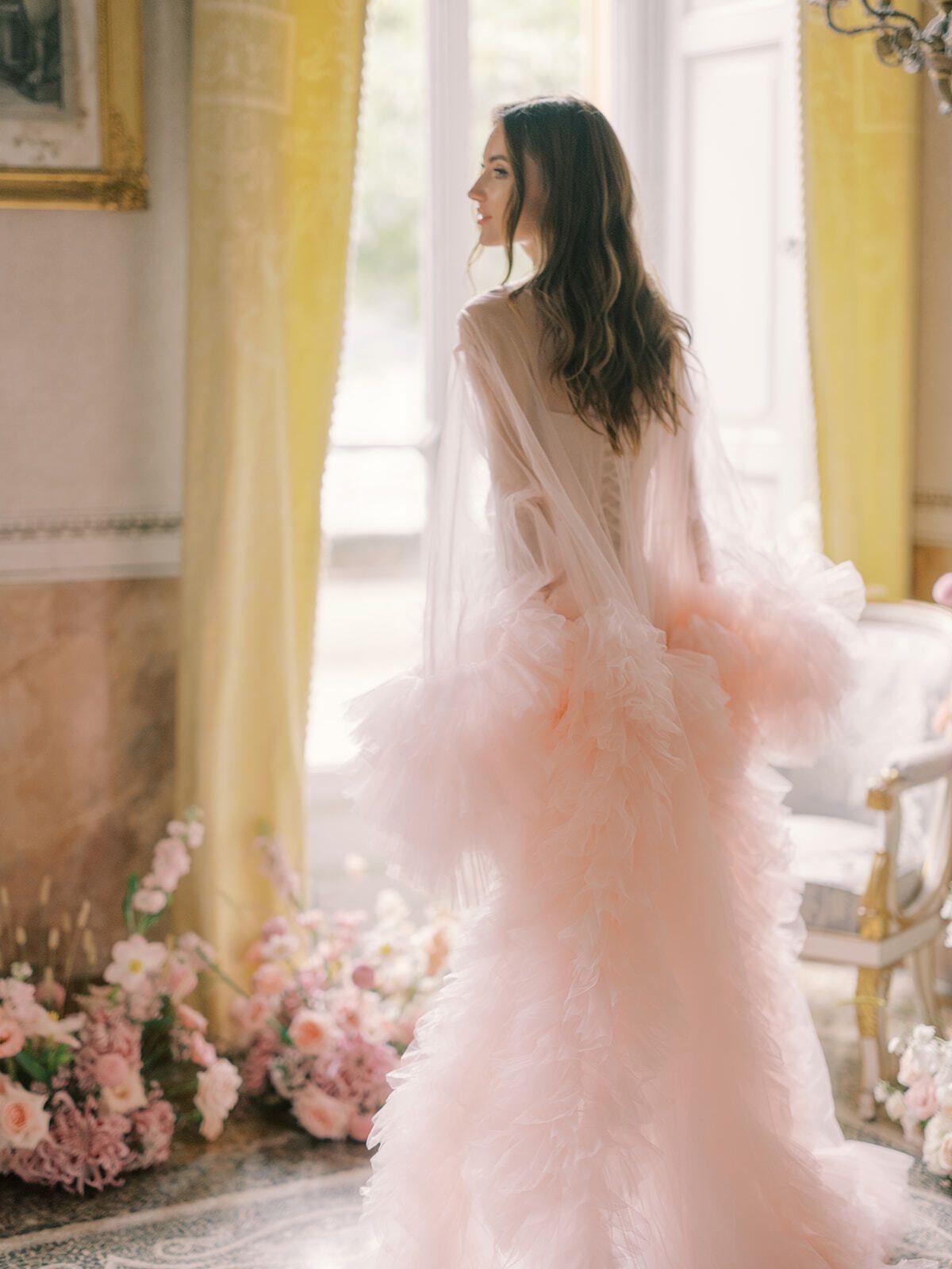 Novia con bata de tul rosa empolvado junto a una ventana soleada