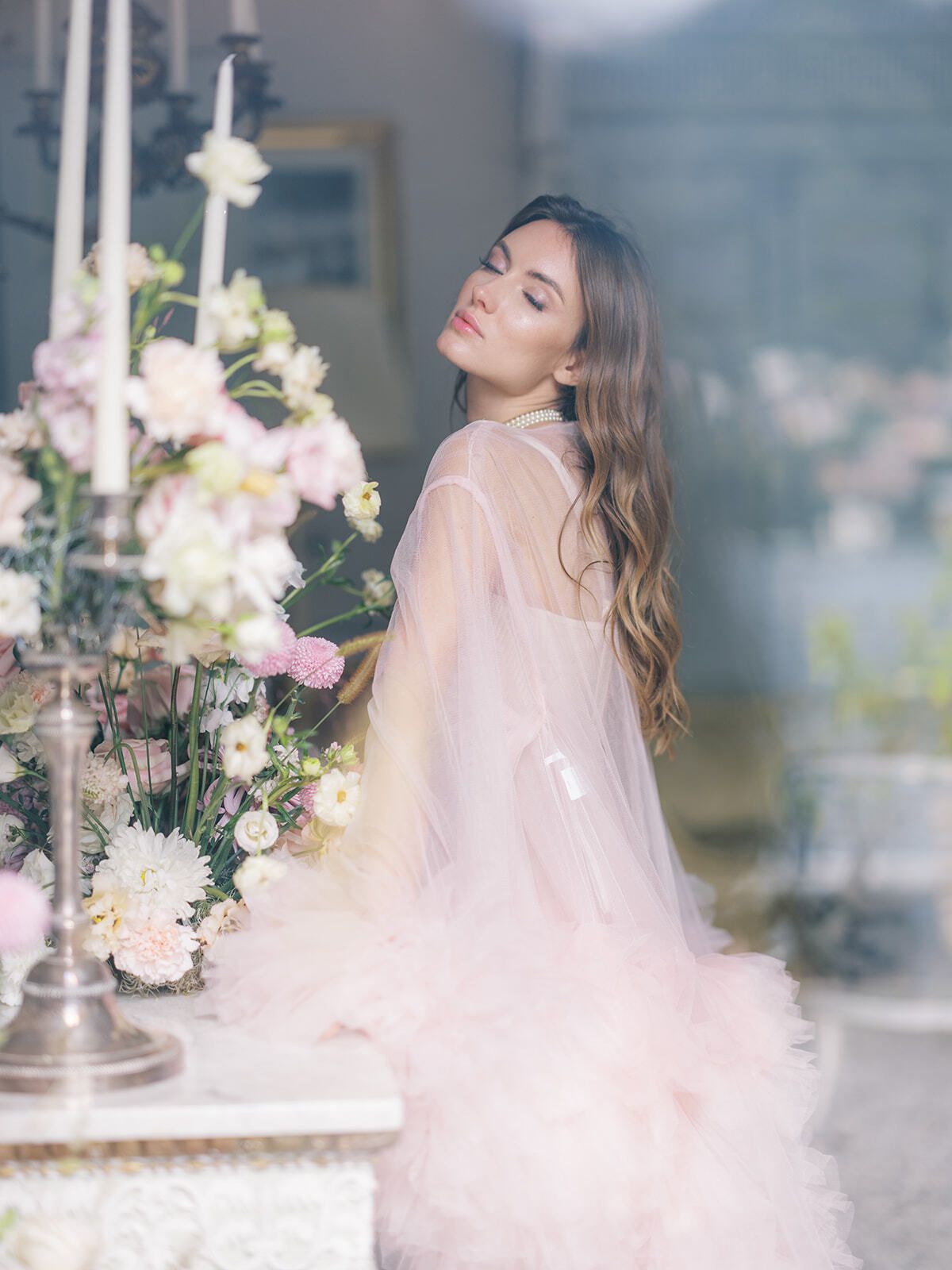 Novia con vestido transparente rosa empolvado entre velas y flores