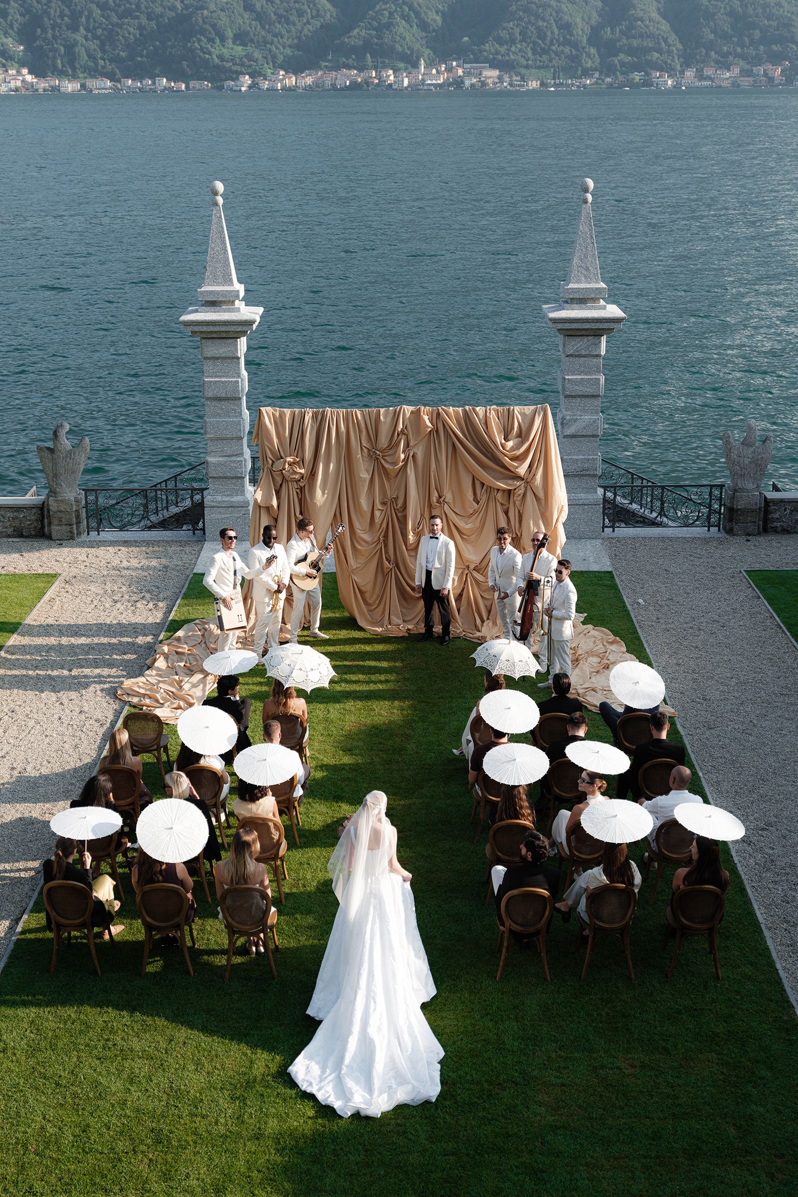 Bride walking up the lakeside lawn to a gold-draped ceremony at Villa Balbiano, guests holding white parasols